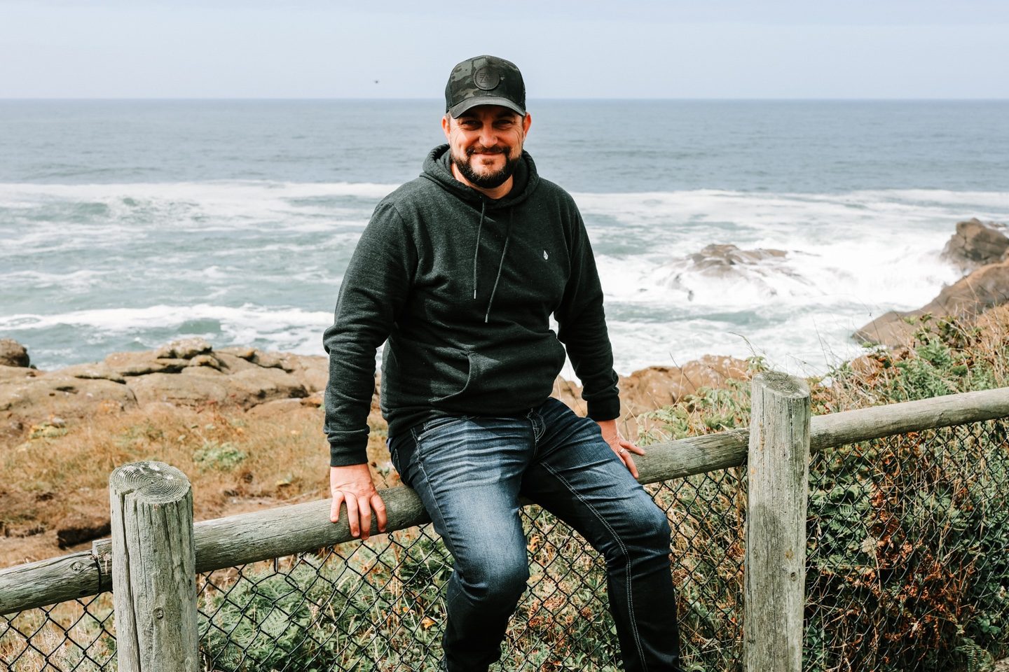 Joshua Brown sitting on a wooden rail above the ocean on the Oregon coast.