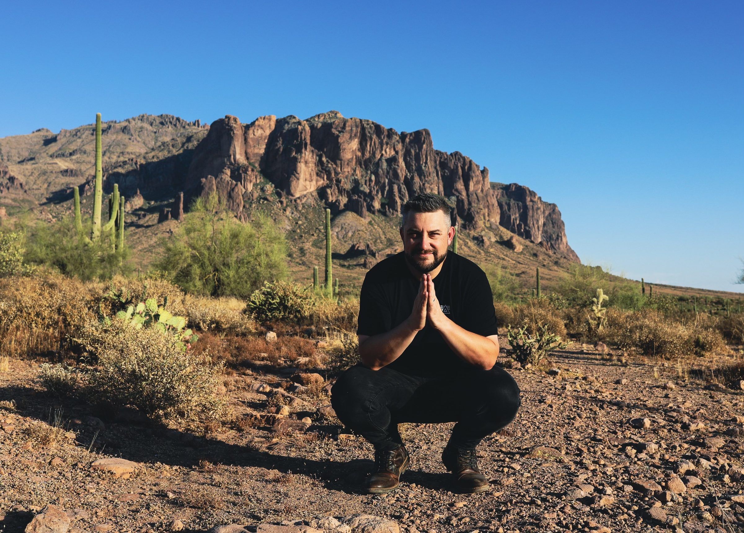 Joshua Brown crouching in the desert with his hands pressed together, saguaro cactus and a rocky mountain behind him.