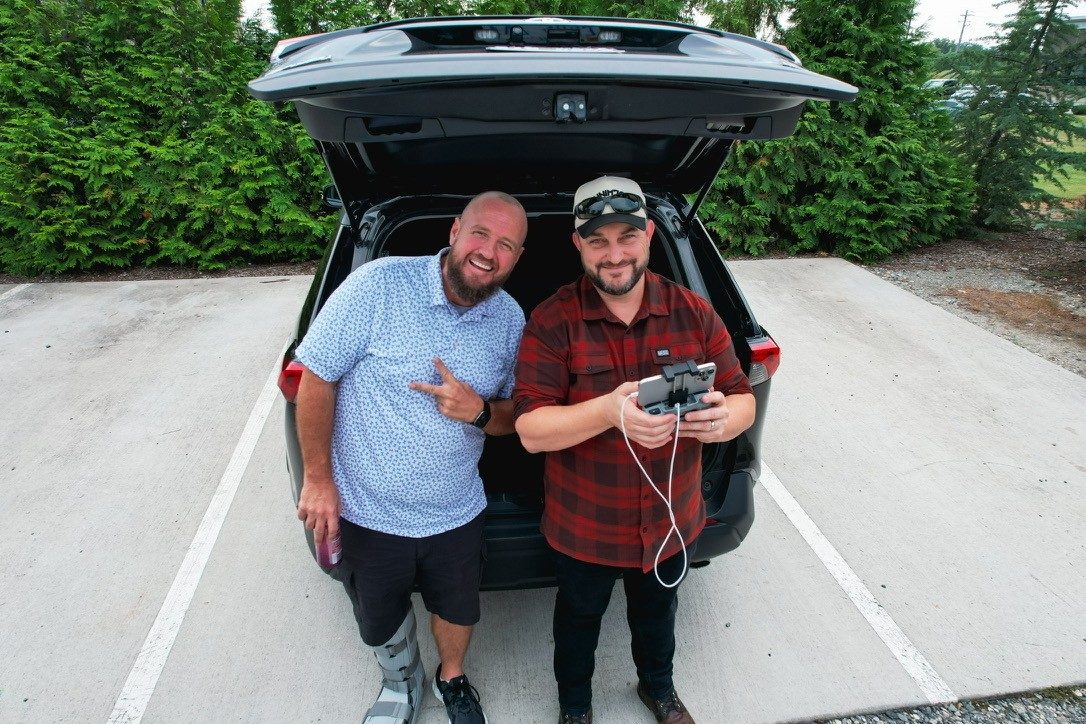 Joshua Brown in a red flannel and cap holding a drone controller beside a friend at an open tailgate.