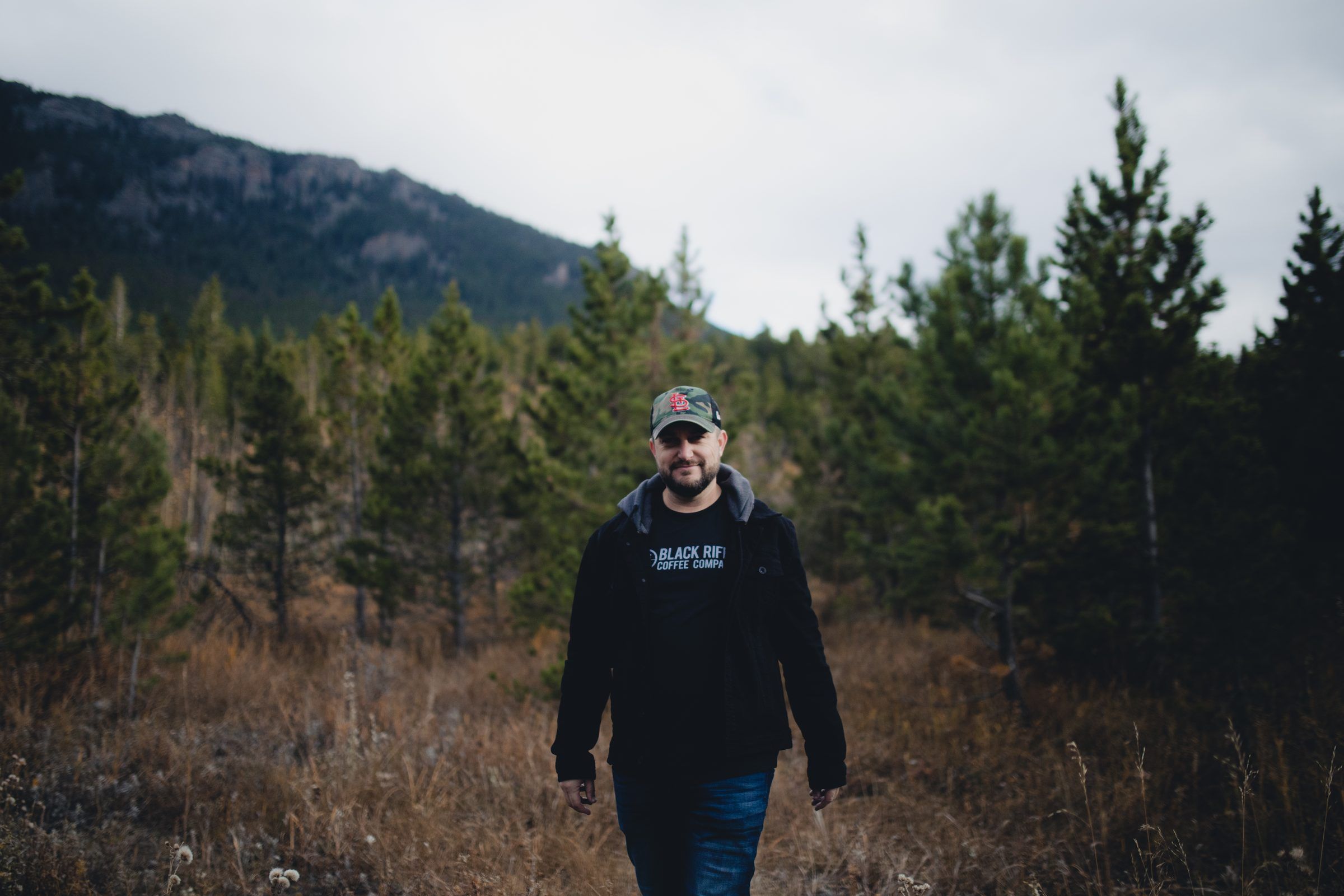 Joshua Brown walking toward the camera through tall grass and pine trees below a mountain.