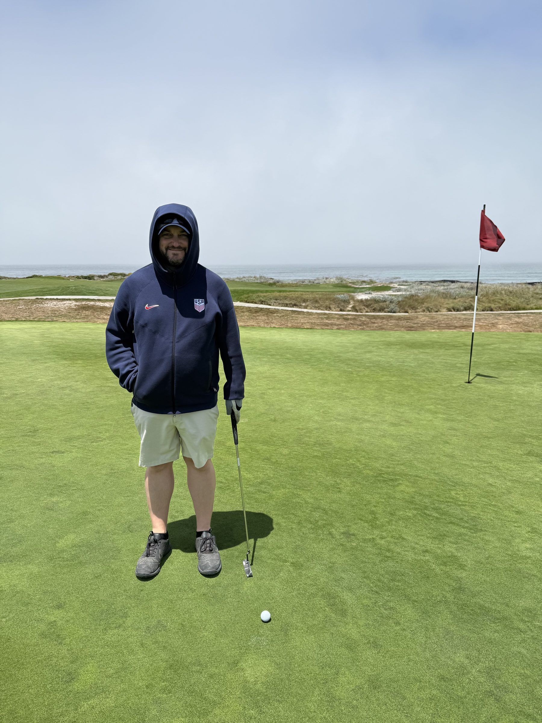 Joshua Brown in a hooded jacket standing on a golf green near the ocean, putter in hand.