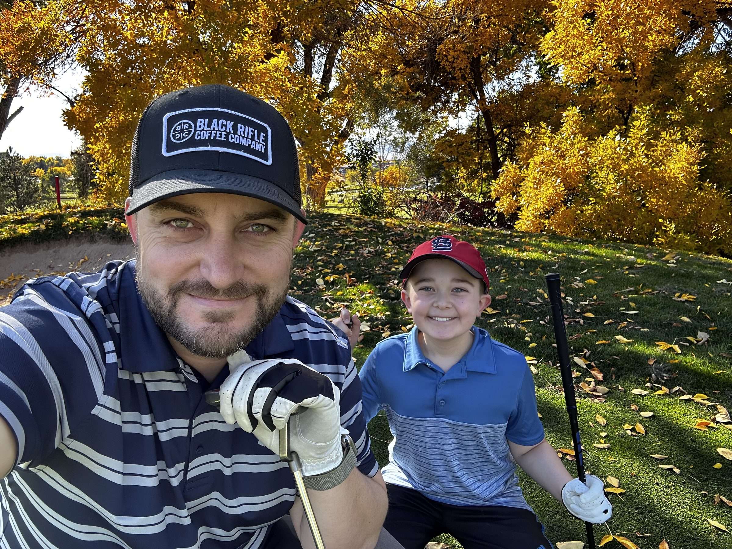 Joshua Brown taking a selfie with his son on a golf course, with bright fall trees behind them.
