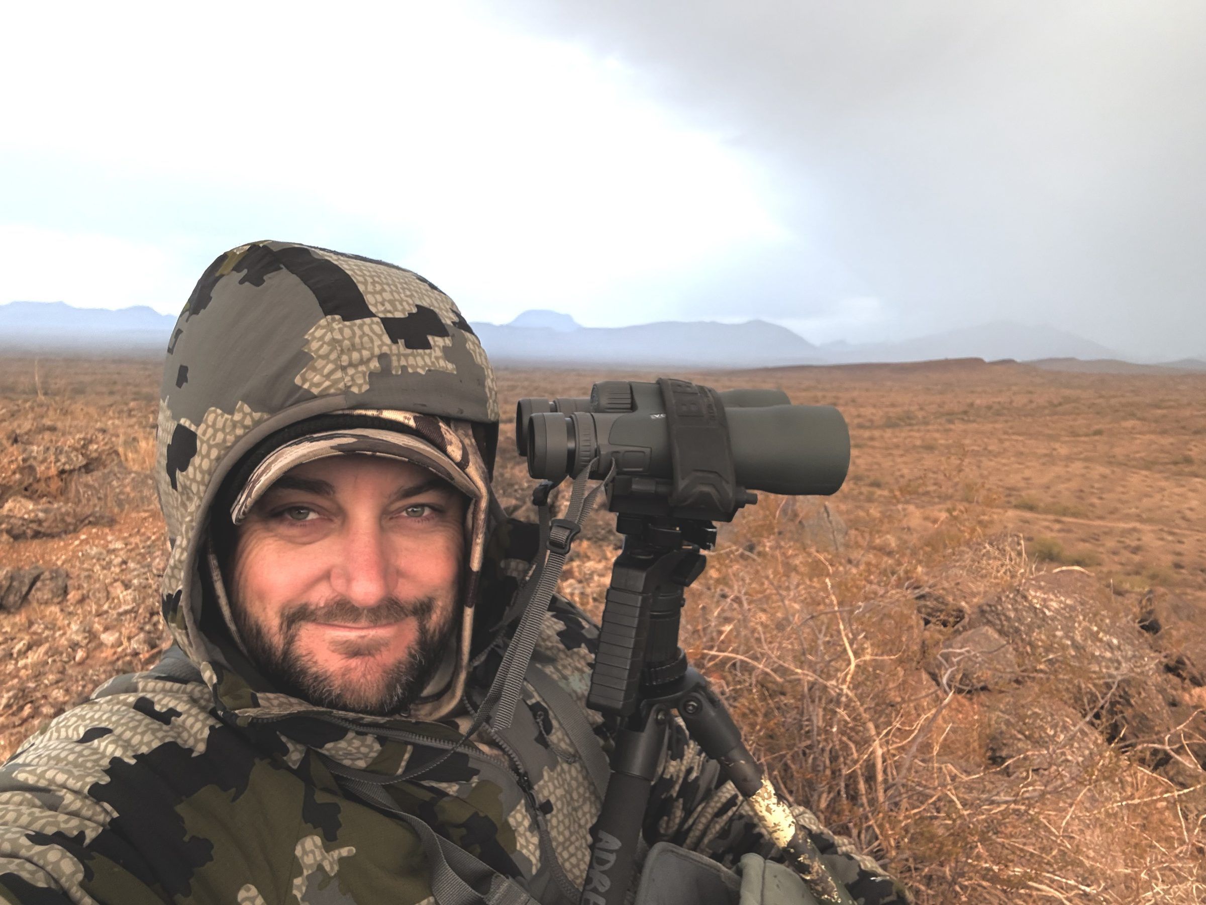 Joshua Brown in camouflage smiling beside binoculars on a tripod in open desert country.
