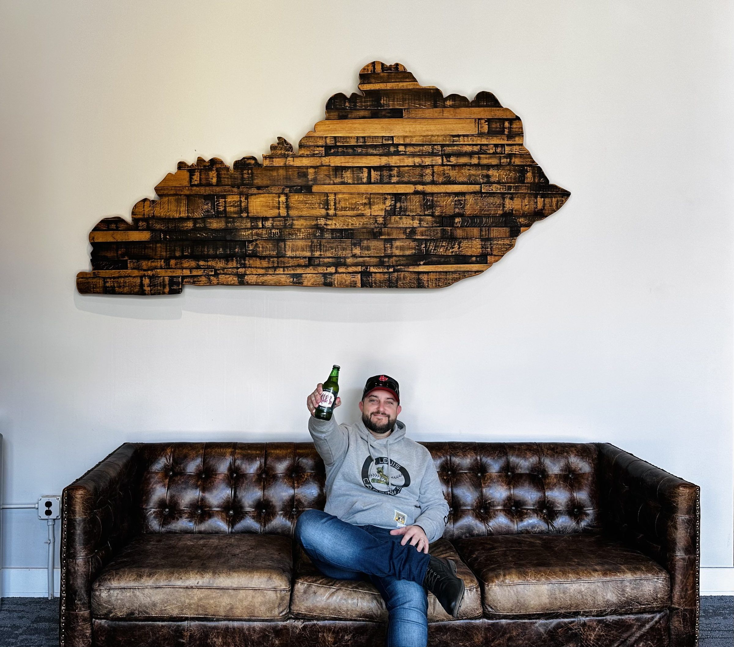 Joshua Brown on a leather couch under a wooden sign shaped like the state of Kentucky, raising a bottle.