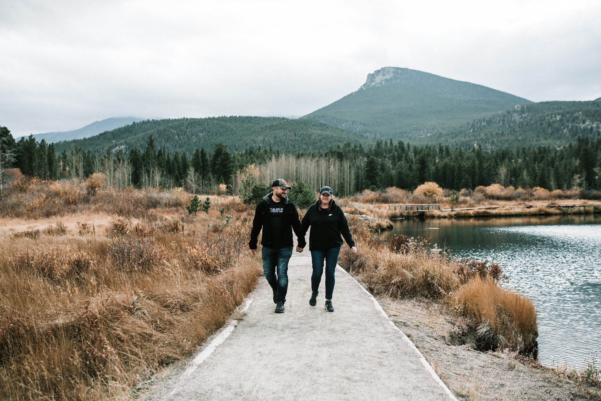Joshua Brown and Laura walking hand in hand on a path beside a mountain lake.