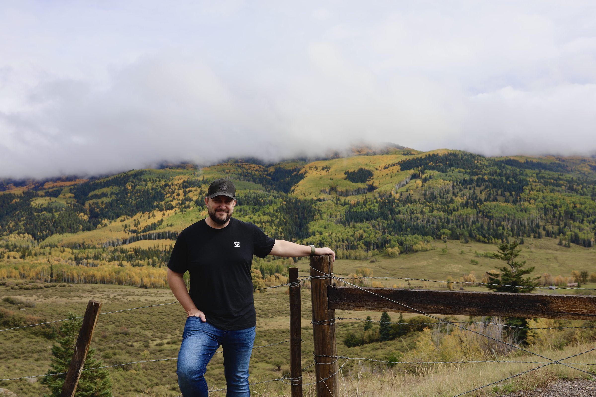 Joshua Brown leaning on a wooden fence post with green and gold mountainsides and low clouds behind him.