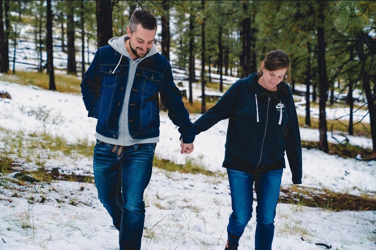 Joshua Brown and Laura walking hand in hand through light snow among pine trees.