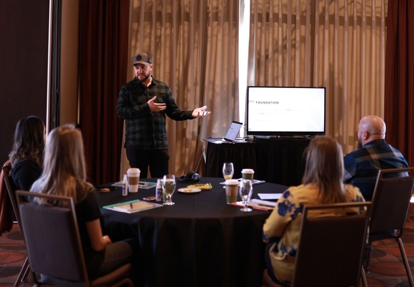 Joshua Brown standing and talking with a small group seated around a table, with a slide on a screen behind him.