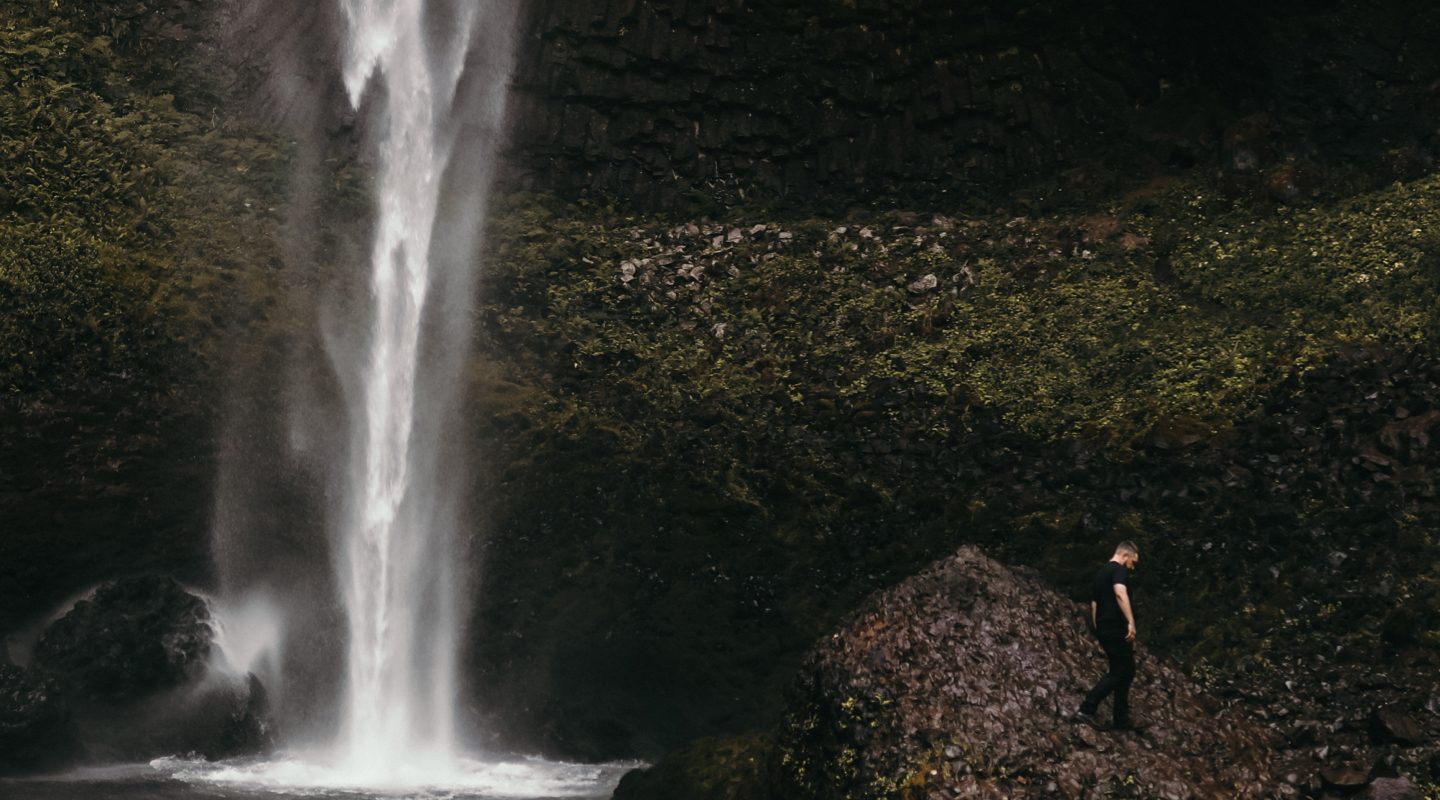 A small figure of Joshua Brown walking on rocks beneath a tall waterfall.